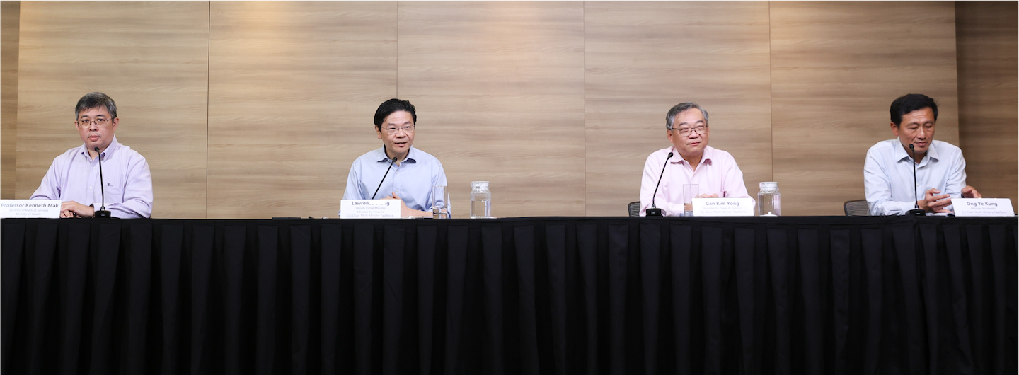 Four men behind a black-draped table, nameplates visible, with microphones, at a press conference.